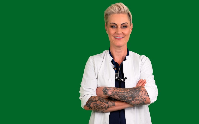 Doc Caro steht mit verschränkten Armen in weißem Arztkittel vor grünem Hintergrund