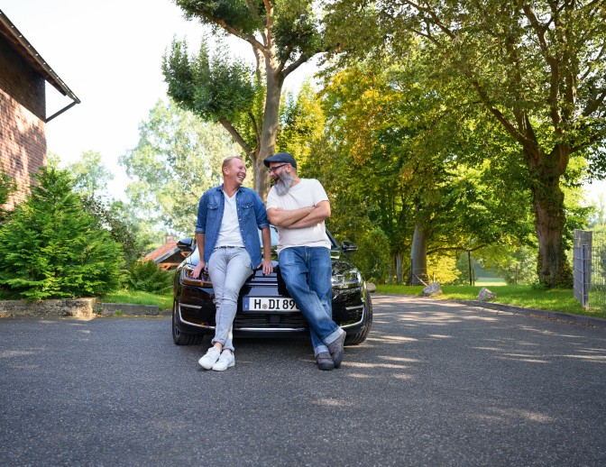 Zwei Männer lehnen entspannt an der Motorhaube eines dunklen Autios und lachen sich an.
