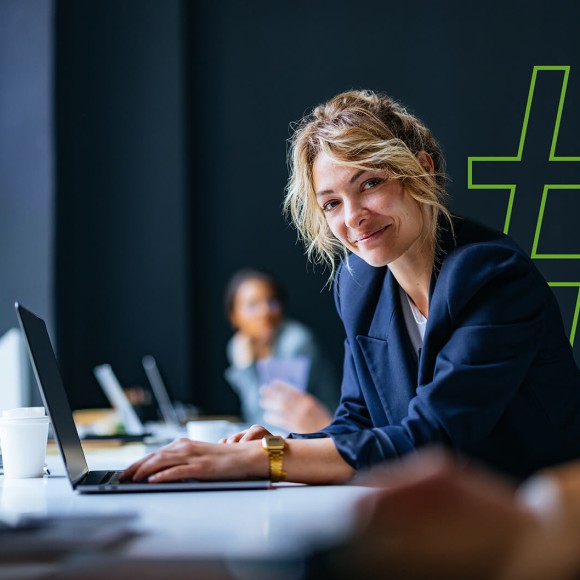 Eine Frau sitzt an einem Schreibtisch und arbeitet an einem Laptop in einem Büro. Im Hintergrund ist ein großes Hashtag-Symbol sichtbar. 
