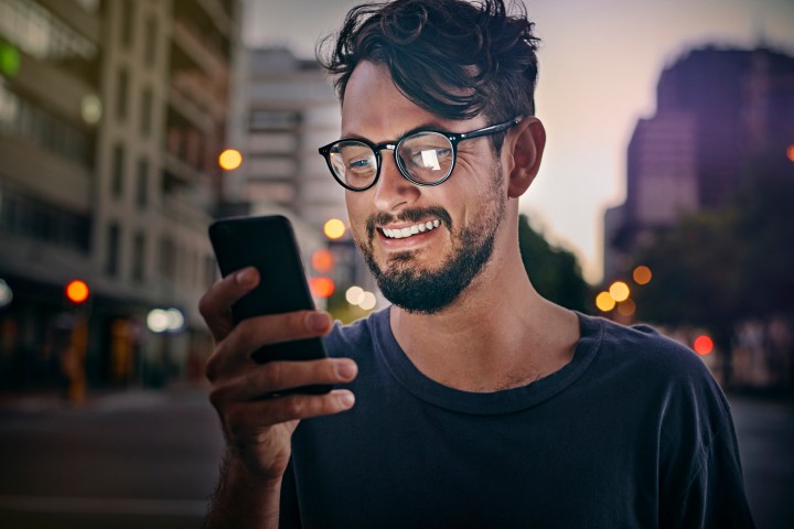 Dunkelhaariger Mann mit Bart und schwarzer Hornbrille schaut lachend auf sein handy, im Hintergrund ist unscharf eine Straße mit Hochhäusern und Laternen zu sehen im Licht der untergehenden Sonne.