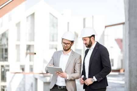 Zwei Architekten auf einem Bauobjekt, die sich unterhalten. Beide Herren tragen einen weißen Bauhelm und einen Anzug.