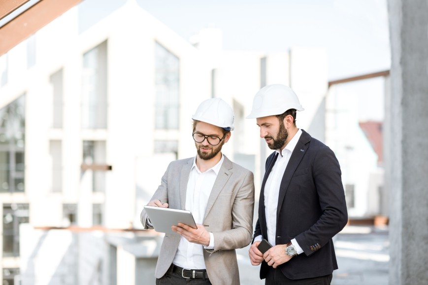 Zwei Architekten auf einem Bauobjekt, die sich unterhalten. Beide Herren tragen einen weißen Bauhelm und einen Anzug.