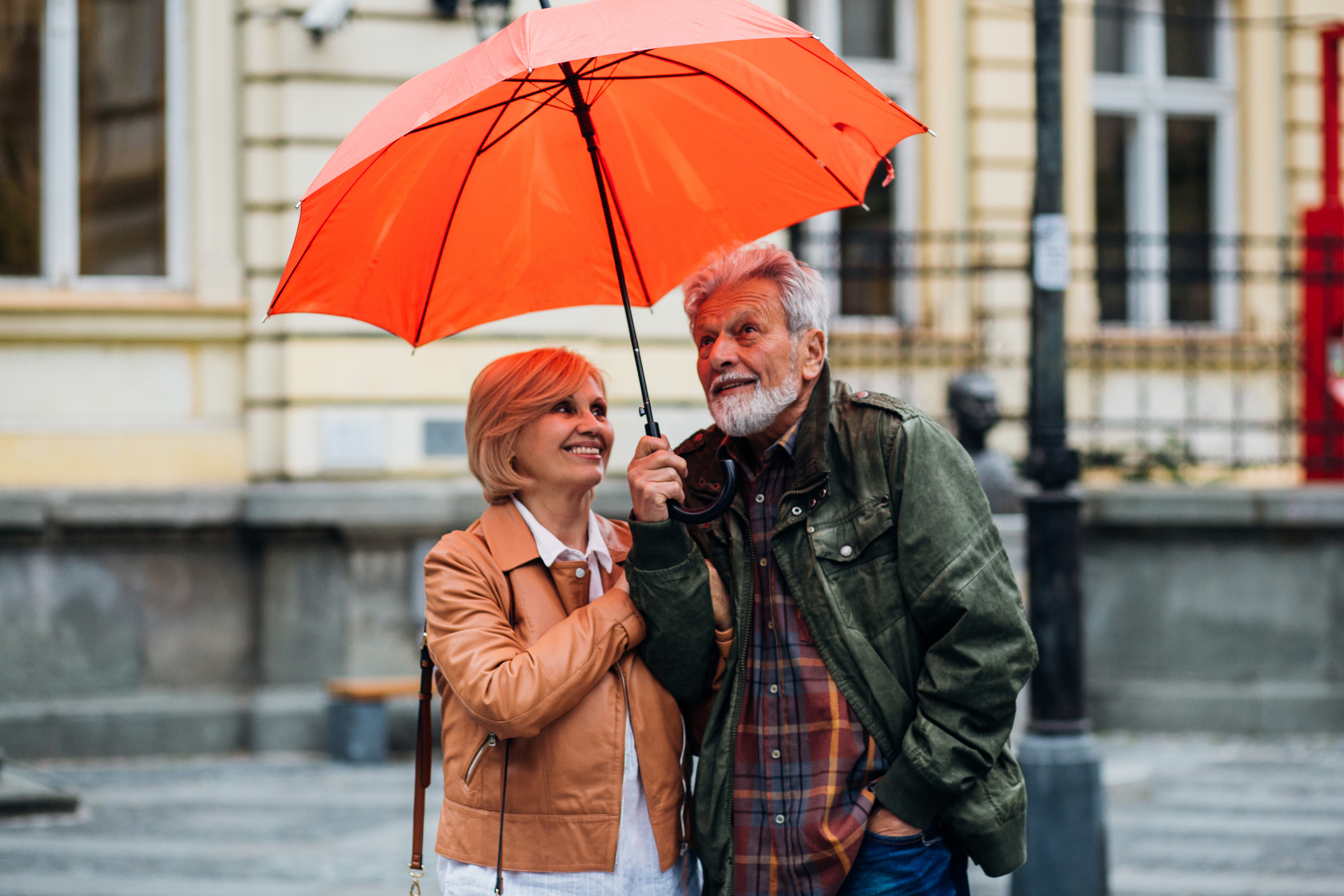 Ein älteres Ehepaar, links eine Frau und rechts ein Mann, stehen unter einem oragngenen regenschrim. der Hintergrund ist unscharf , jedoch erkennt man ein gelbes Haus und vor dem Haus ist eine bank und rechts ist ein Zaun und vor dem Zaun steht ein Laternemast.