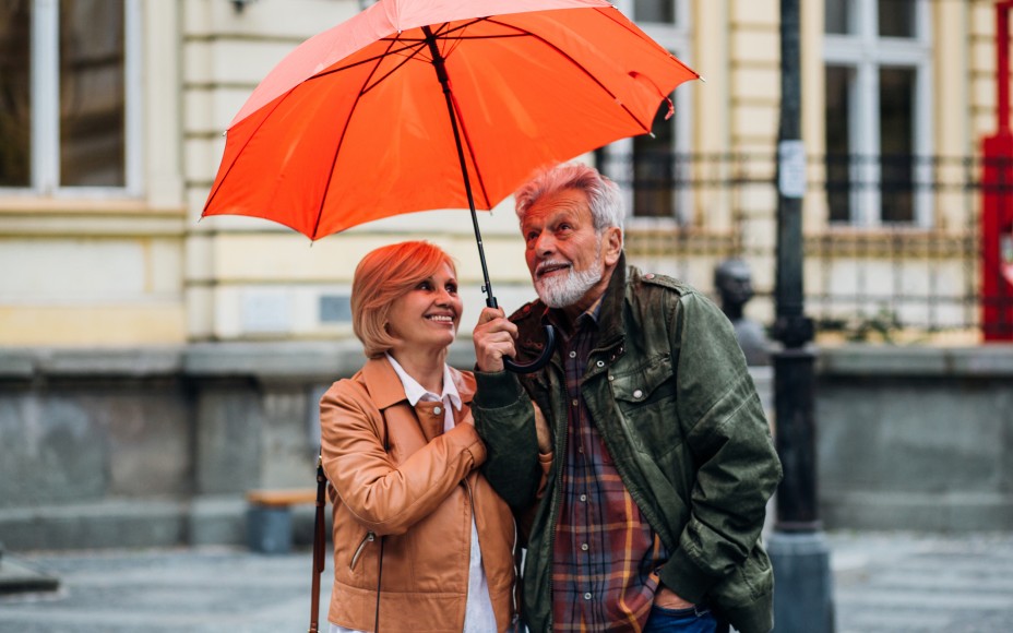 Ein älteres Ehepaar, links eine Frau und rechts ein Mann, stehen unter einem oragngenen regenschrim. der Hintergrund ist unscharf , jedoch erkennt man ein gelbes Haus und vor dem Haus ist eine bank und rechts ist ein Zaun und vor dem Zaun steht ein Laternemast.