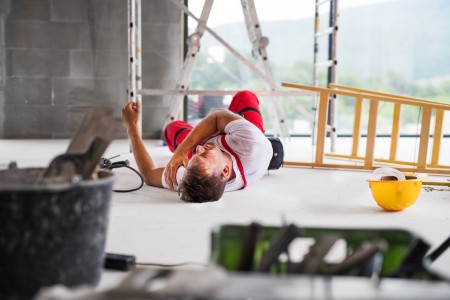 Mann in Arbeitshose leigt auf Boden. Man sieht eine umgekippte Leiter, neben ihm liegt ein gelber Schutzhelm, im Hintergrund ist eine weitere Leiter zu erkennen und ein Gerüst, im Vordergrund stehen andere Arbeitsutensilien