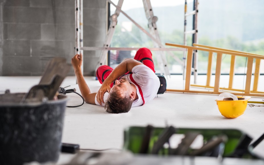 Mann in Arbeitshose leigt auf Boden. Man sieht eine umgekippte Leiter, neben ihm liegt ein gelber Schutzhelm, im Hintergrund ist eine weitere Leiter zu erkennen und ein Gerüst, im Vordergrund stehen andere Arbeitsutensilien