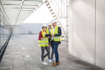 Drei Menschen haben gelbe Westen und weiße Helme an. Eine Frau, die lächelnd in die Kamera guckt, steht in der Links und hat ein Ipad in der Hand. Rechts von ihr stehen zwei Männer, die auf das Ipad gucken und zeigen.