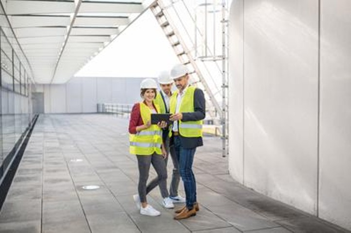 Drei Menschen haben gelbe Westen und weiße Helme an. Eine Frau, die lächelnd in die Kamera guckt, steht in der Links und hat ein Ipad in der Hand. Rechts von ihr stehen zwei Männer, die auf das Ipad gucken und zeigen.