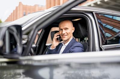 Ein Mann sitzt in einem schwarzem Auto und wird durch die runtergelassene Fensterscheibe fotografiert. Er guckt lächelnd in die Kamera und hält sein Telefon ans Ohr.