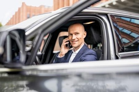 Ein Mann sitzt in einem schwarzem Auto und wird durch die runtergelassene Fensterscheibe fotografiert. Er guckt lächelnd in die Kamera und hält sein Telefon ans Ohr.