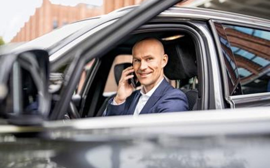 Ein Mann sitzt in einem schwarzem Auto und wird durch die runtergelassene Fensterscheibe fotografiert. Er guckt lächelnd in die Kamera und hält sein Telefon ans Ohr.