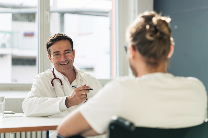 Auf der einen Seite mit dem Bilck zum Patienten sitzt ein Arzt mit einem Stift in der Hand und gegenüber von dem Arzt mit dem Rücken zur Kamera sitzt der Patient im Rollstuhl.