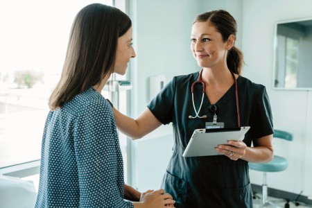 Links ist eine Patienten, die auf einer Liege sitzt und auf der rechten Seite steht eine Ärztin mit grünen Kittel. In der linken Hand hält sie ein Brett mit Zettel und mit die rechte Hand liegt auf der Schulter, der Patienten.