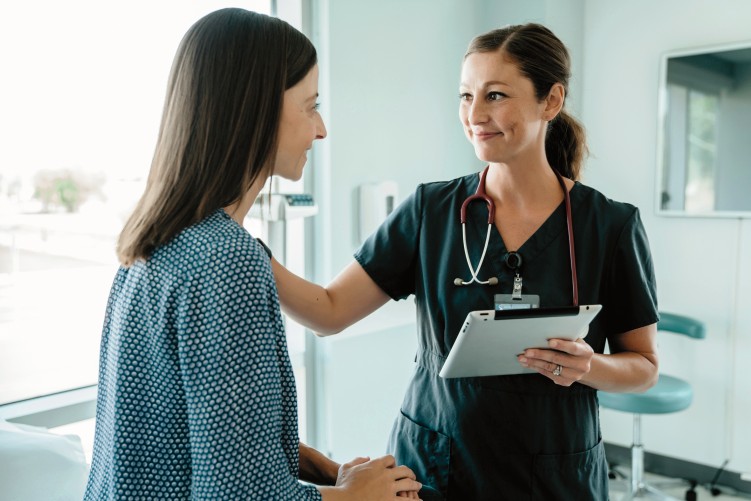 Links ist eine Patienten, die auf einer Liege sitzt und auf der rechten Seite steht eine Ärztin mit grünen Kittel. In der linken Hand hält sie ein Brett mit Zettel und mit die rechte Hand liegt auf der Schulter, der Patienten.
