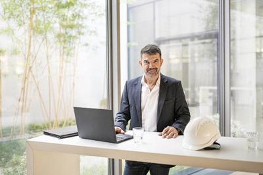 Ein Mann steht an einem Schreibtisch und lächelt in die Kamera. Vor ihr auf dem Schreibtisch steht ein PC, ein Helm, ein Handy, ein Ipad und ein Glas mit durchsichtiger Flüssigkeit.