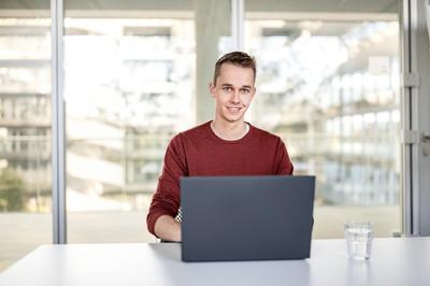 Ein Mann, der in die Kamera guckt, sitzt an einem Schreibtisch und hat vor sich ein Laptopn an dem er arbeitet.