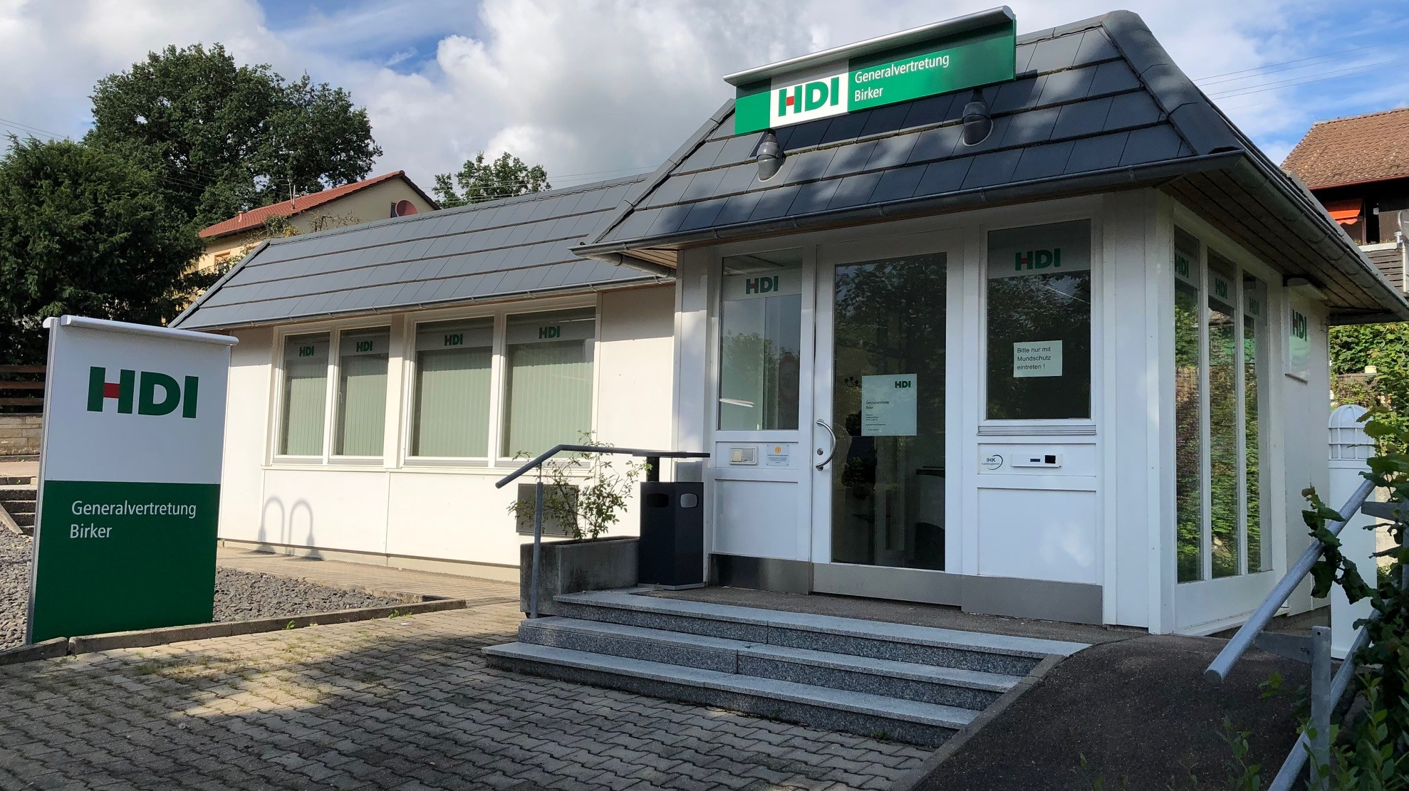 Man sieht die Agentur von außen. Es ist ein weißes Gebäude mit schwarzem Dach. Auf dem Dach und em Parkplatz befinden sich Schilder mit dem HDI Logo.