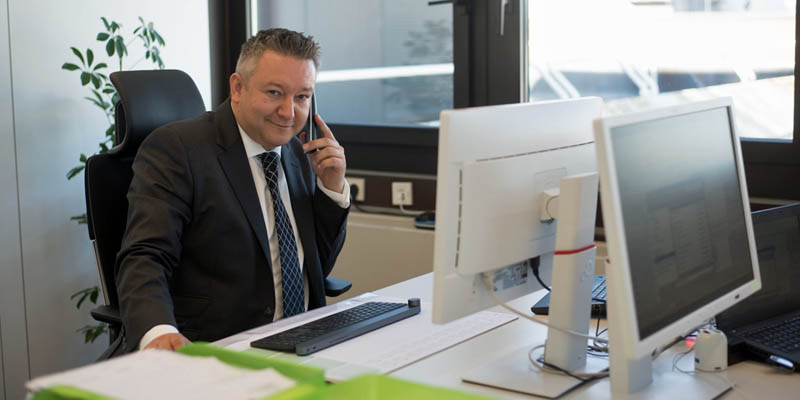Markovic sitzt auf einem schwarzen Stuhl an einem Schreibtsich und ist am telefonieren, dabei lächelt er in die Kamera. Hinter Ihm ist ein Fenster und links daneben steht eine grüne Pflanze, die durch den Stuhl verdeckt wird.