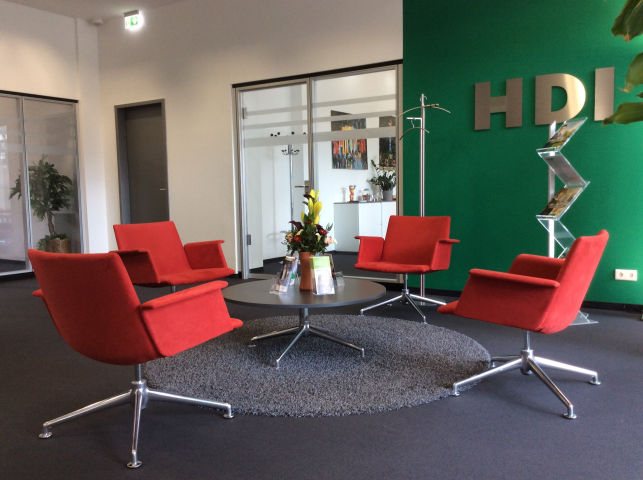 In der Mitte steht ein runder Glastisch, auf dem Blumen und Flyer stehen. Um den runden Glastisch stehen vier rote Stühle. Im Hintergrund ist eine Glastür, die zu einem weiteren Büro führt. Rechts ist eine grüne Wand an dem silber HDI Bustaben hängen.