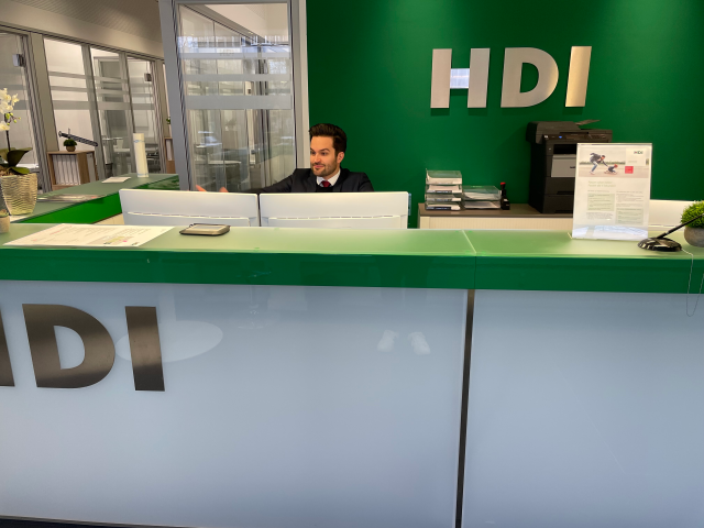 weiß, grüne Theke, hinter der ein Mitarbeiter vor zwei Bildschirmen sitzt. Links hinter Ihm sind Glasfenster, die Büros zeigen. Rechts von Ihm ist ein braun, weißes regal zusehen auf dem drucker und Fächer stehen. Über dem Regal an der grünen Wand hängt das silberne HDI Logo.