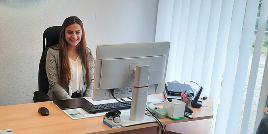 Man sieht die Mitarbeiterin Agnesa Hoti am Schreibtisch sitzen. Vor sich hat sie einen PC-Bildschirm und -Zubehör zu stehen sowie Büroartikel. Sie schaut lächelnd in die Kamera.