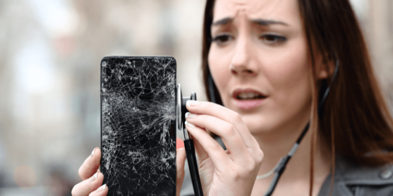 Eine Frau mit geschocktem Gesicht hält ein Handy mit zersprungenem Bildschirm in der Hand.