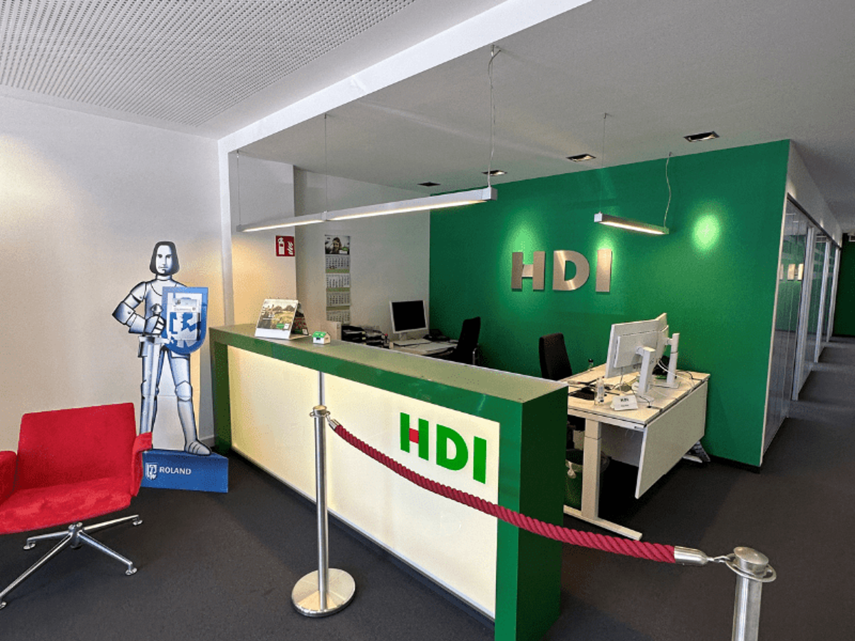Man erkennt eine weiß, grüne Theke. Vor der Theke steht ein roter Stuhl, ein rotes Absperrband und eine Ritter Papier Startue. Hinter der Theke sind Tische und Stühle. An der der Wand hägt das HDI Logo