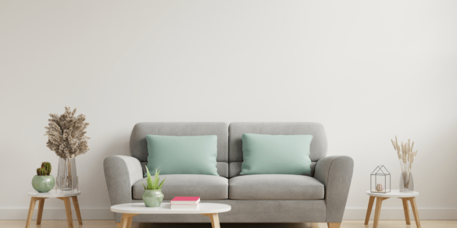 Das Sofa ist hellgrau und vor dem Sofa steht ein kleiner weißer Tisch, auf dem eine Pflanze und ein Buch drauf liegen.