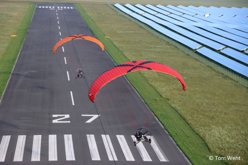 Zwei Motorgleitschirme im Landanflug auf eine Fluglandebahn. Ein Gleitschirm ist orange, der ander ist rot. Die Landebahn ist umgeben von grünem Feld und einem Feld mit Solaranlagen befindet sich in der Nähe.