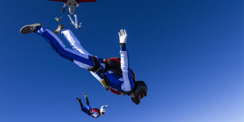Mehrere Skydiver kurz nach Sprung aus Flugzeug. Die Skydiver tragen weiß-blaue Anzüge und Helme. Der Himmel ist strahlend blau und wolkenlos.