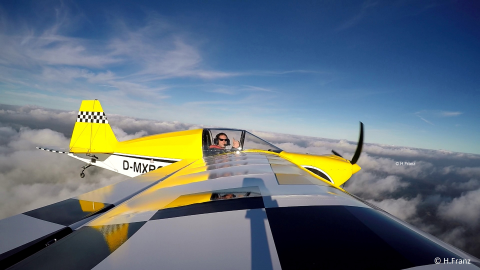 Ultraleichtpilot in Flugzeug in der Luft. Das Flugzeug ist größtenteils gelb. Der Pilot zeigt einen Daumen hoch in die Kamera. Der Himmel ist teilweise bewölkt.