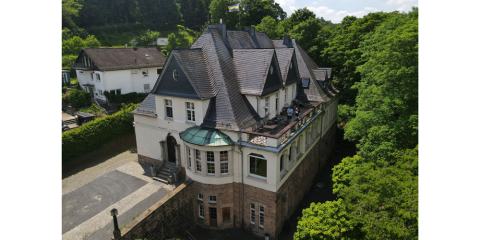 das Verbindungshaus ist weiß und hat ein graues Dach, links neben dem Haus steht ein weiteres Haus und überall sind grüne Bäume