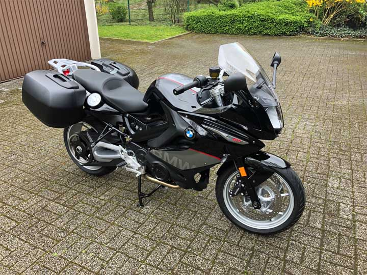 ein schwarzes Motorrad steht auf Steinen vor einer braunen Garage. Im Hintergrund ist ein grüne Wiese und grüne Büsche
