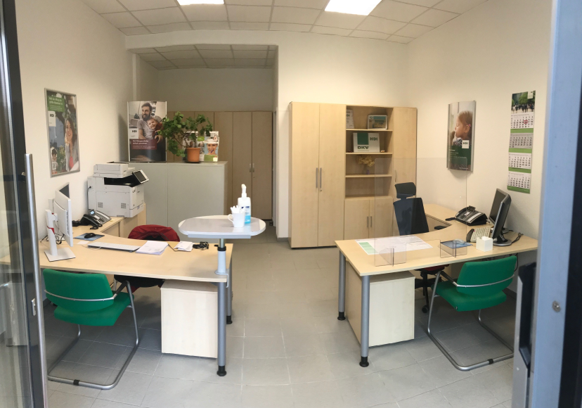 Links und Rechts stehen Tische mit grünen Stühlen und shcwarzen Stühlen. Auf beiden Tischen stehen PC. Hinter dem rechten Tisch steht ein großes braunes Regal. Und an den Wänden hängen HDI Plakate. Zwischen den Tischen ist ein Durchgang,