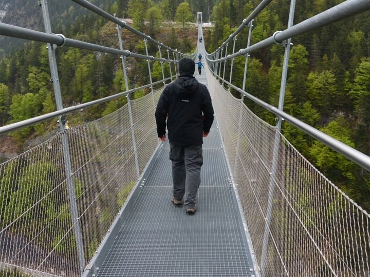 Ein Mann läuft über eine Stahlbrücke, vor ihm sind vier weiter Personen in weiter ferne, neben de Brücke sind viele grüne Bäume