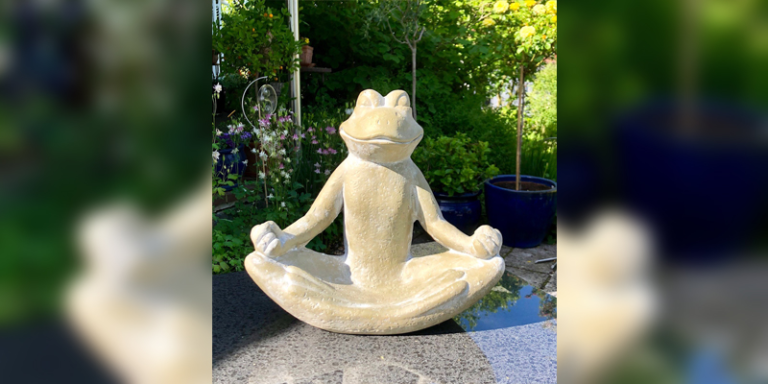 meditierter Frosch im Schneidersitz sitzt auf einem Steintisch in einem Garten, hinter ihm sind weiße und lilane Blumen und grüne Bäume