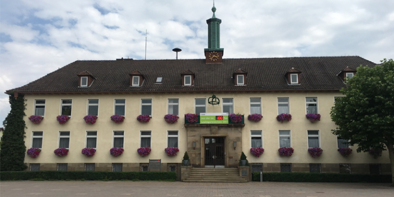 gelbes Haus mit braunen Dach, blauen Himmel und rechts neben dem Haus ein grüner Baum