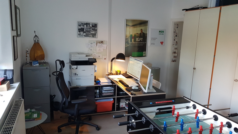 in der Mitte an der Wand steht ein Schreibtisch mit PC und Lampe, davor steht ein Stuhl, neben dem Stuhl ein Drucker und dahinter ein Schrank. Vor dem Schreibtisch stehen zwei große Schränke und neben dem Schreibtisch steht ein Tischkicker