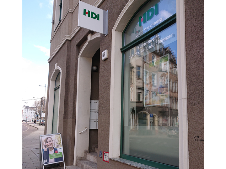 Rechts auf der Seite ist die Agentur, die in einem roten Gebäude ist mit einem großen Fenster. Vor der Agentur steht ein HDI Aufsteller und an dem Gebäude hängt das HDI Schild.