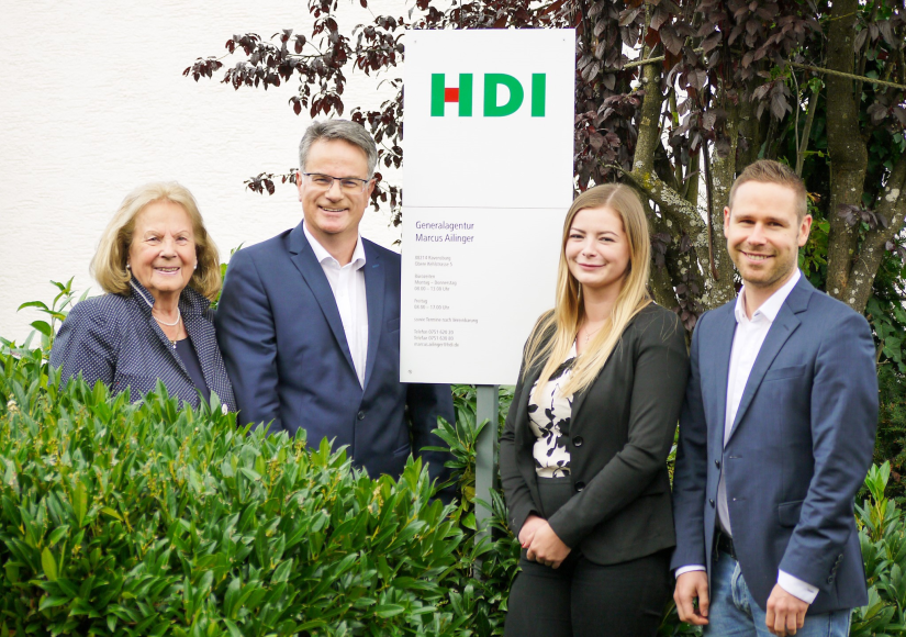 Ein Teamfoto im Garten neben dem HDI Eingangsschild zur Generalagentur Marcus Ailinger. Links vom Schild Herr Ailinger und Frau AIlinger, rechts vom Schild zwei Teammitglieder aus der Agentur. Alle Personen tragen ein förmliches Outfit und lächeln in die Kamera.
