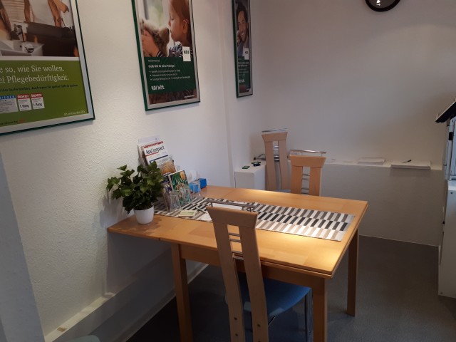 Ein Holztisch und zwei Stühle, an der Wand hängen Plakate mit HDI Werbun. Auf dem Tisch stehen Flyer und andere Werbungen sowie eine Grünpflanze.