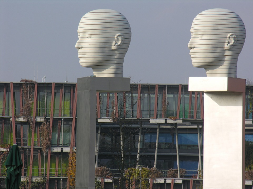Zwei Statuen in Form von weißen Köpfen auf Podesten, im Hintergrund sieht man ein lange, braunes Gebäude mit vielen Fenstern