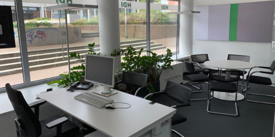 Man sieht ein Büro, im Vordergrund befindet sich ein Schreibtisch mit Stuhl, auf dem Schreibtisch steht ein PC und PC-Zubehör sowie ein Telefon, vor der Fensterfront befindet sich eine Bank und Säulen, von denen eine von Zimmerpflanzen umstellt ist, im hinteren Teil des Raums befindet sich ein runder Tisch mit fünf Stühlen