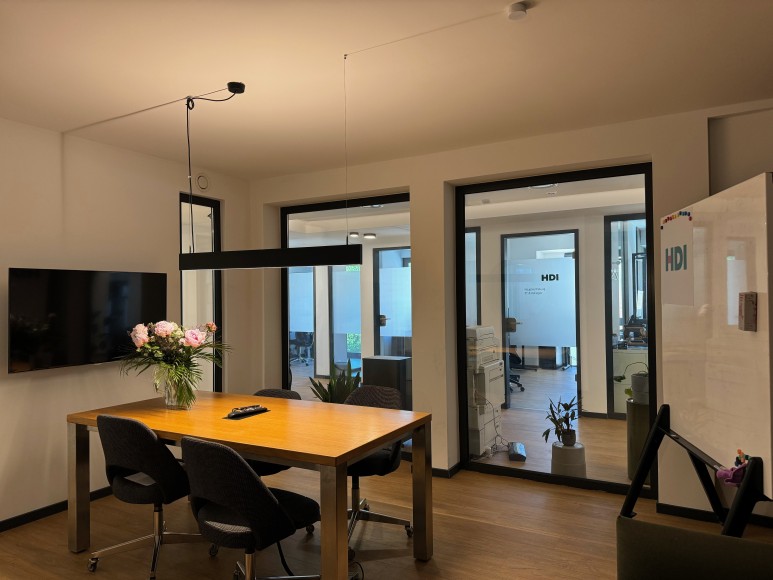 In der Mitte steht ein brauner Tisch, um den Tisch stehen vier schwarze Stühle und auf dem Tisch steht ein Blumestrauß. Links vom Tisch ist ein Fernseher an der Wand und hinter dem Tisch ist eine Fensterfront, durch die man in den Flur gucken kann. Rechts neben dem Tisch steht ein Whiteboard.