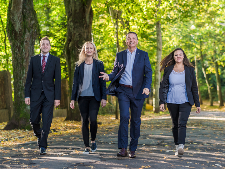 zwei Mitarbeiter und zwei Mitarbeiterinnen, die gehen. sie gehen auf einem grauen gehweg und neben dem Gehweg und hinter dem Mitarbeitern stehen grüne Bäume mit brauen Bäumstämmen