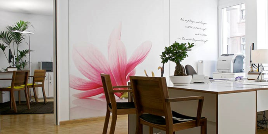 Büro mit einem Tisch und Stühlen und einer weißen Tapete auf der eine rosa Blume ist. Links ist ein Durchgang ,der in ein weiteres Büro führt, indem ein Tisch, zwei Stühle, eine Lampe und eine Pflanze steht.