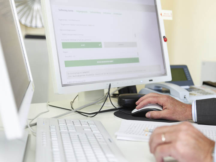 Man sieht einen Schreibtsich mit zwei Monitoren, PC-Zubehör und Büroartikeln. Man sieht zwei Hände von denen eine die Maus in der Hand hält.
