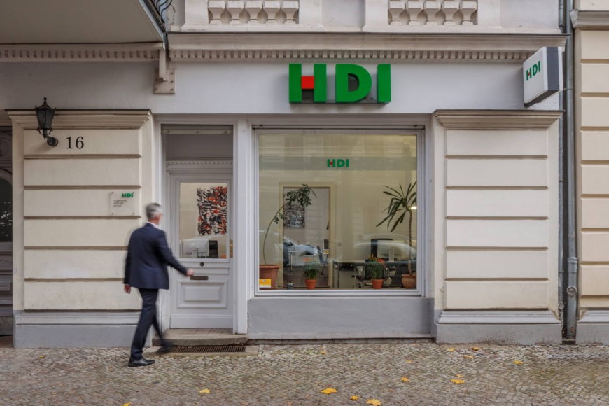 Die Agentur befindet sich in einem beigen Haus. Man erkennt ein fenster, durch dieses sieht man ins Büro. Über diesem Fenster befindet sich das HDI Logo und links neben dem Fenster ist eine Glatür, woe gerade ein Mann drauf zu läuft. Vor der Agentur ist ein grauer Gehweg.