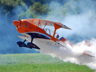 Orangener Modellflieger hebt ab. Hinter dem Flieger ist eine Rauchwolke und der Flieger startet auf einer Wiese. Hinter dem Flieger ist ein Wald mit grünen Bäumen.