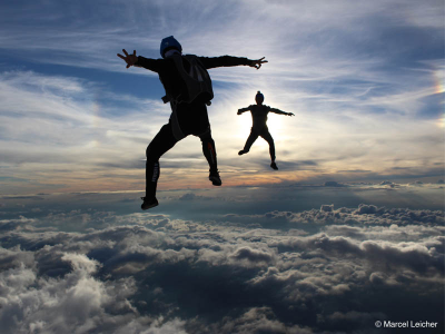 Zwei Menschen sind in der Luft mit Rucksäcken, wo Gleitschirme drin sind. Unter ihnen sind weiße Wolken und über ihnen ist ein blauer Himmel mit einem Schleier weißer Wolken.